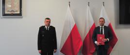 Pomorski komendant wojewódzki PSP wspólnie z prezesem Wojewódzkiego Funduszu Ochrony Środowiska i Gospodarki Wodnej w Gdańsku trzymającego dokument stoją na tle flagi państwowej.