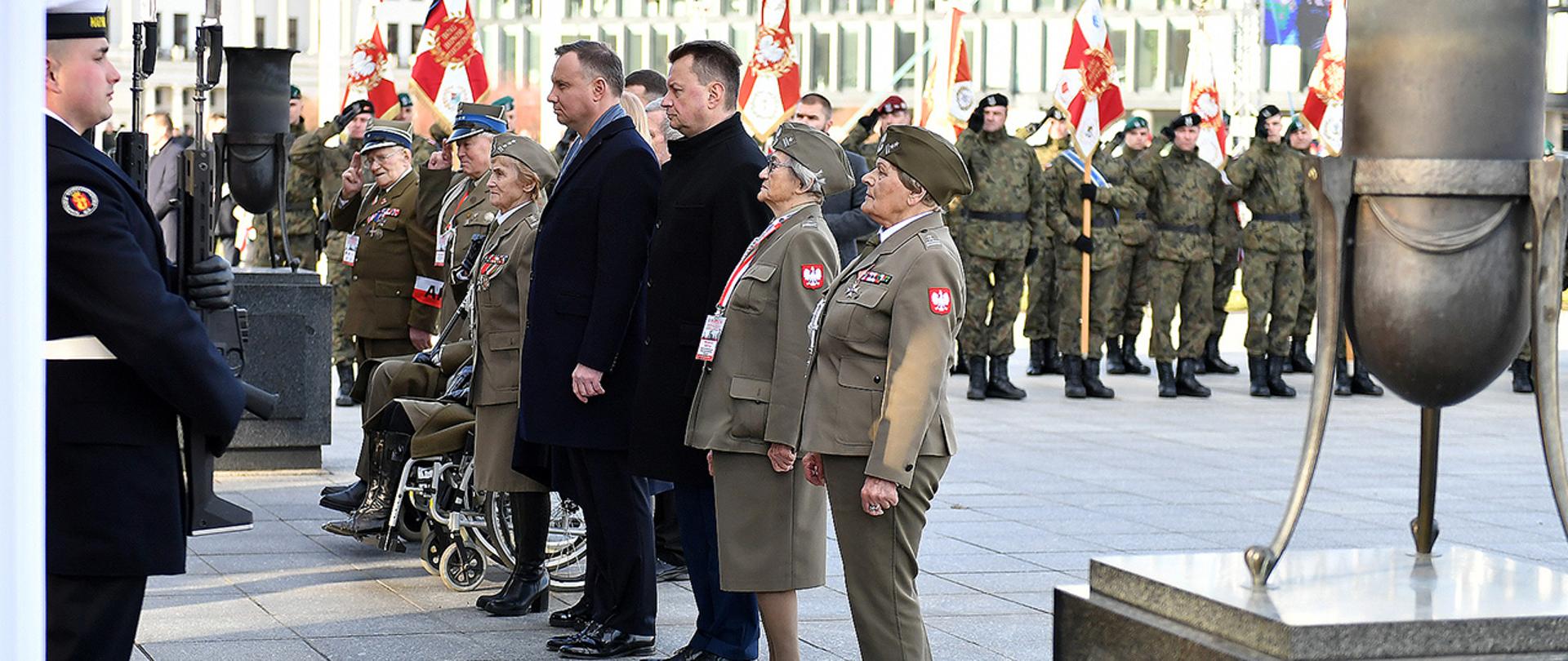 Prezydent RP, szef MON i kombatanci stoją przed Grobem Nieznanego Żołnierza podczas ceremonii złożenia wieńców. W tle żołnierze Wojska Polskiego z pocztami sztandarowymi.
