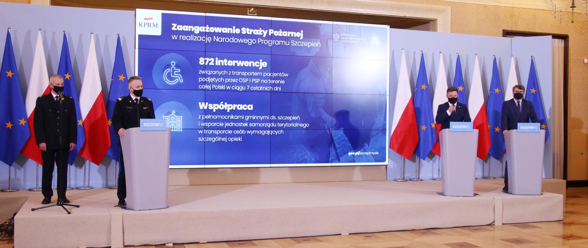 Komendant Główny PSP wraz z przedstawicielami rządu na tle bannera akcji Narodowego Programu Szczepień.