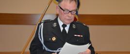 Zebranie sprawozdawcze OSP Śląskowo. Świetlica wiejska w Śląskowie. Prezes zarządu OSP w mundurze galowym stoi za stołem, na tle sceny i sztandaru, przedstawiając porządek zebrania.