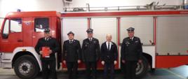 czterech strażaków w ciemnych mundurach i w rogatywkach stoi w szeregu przed czerwonym samochodem strażackim