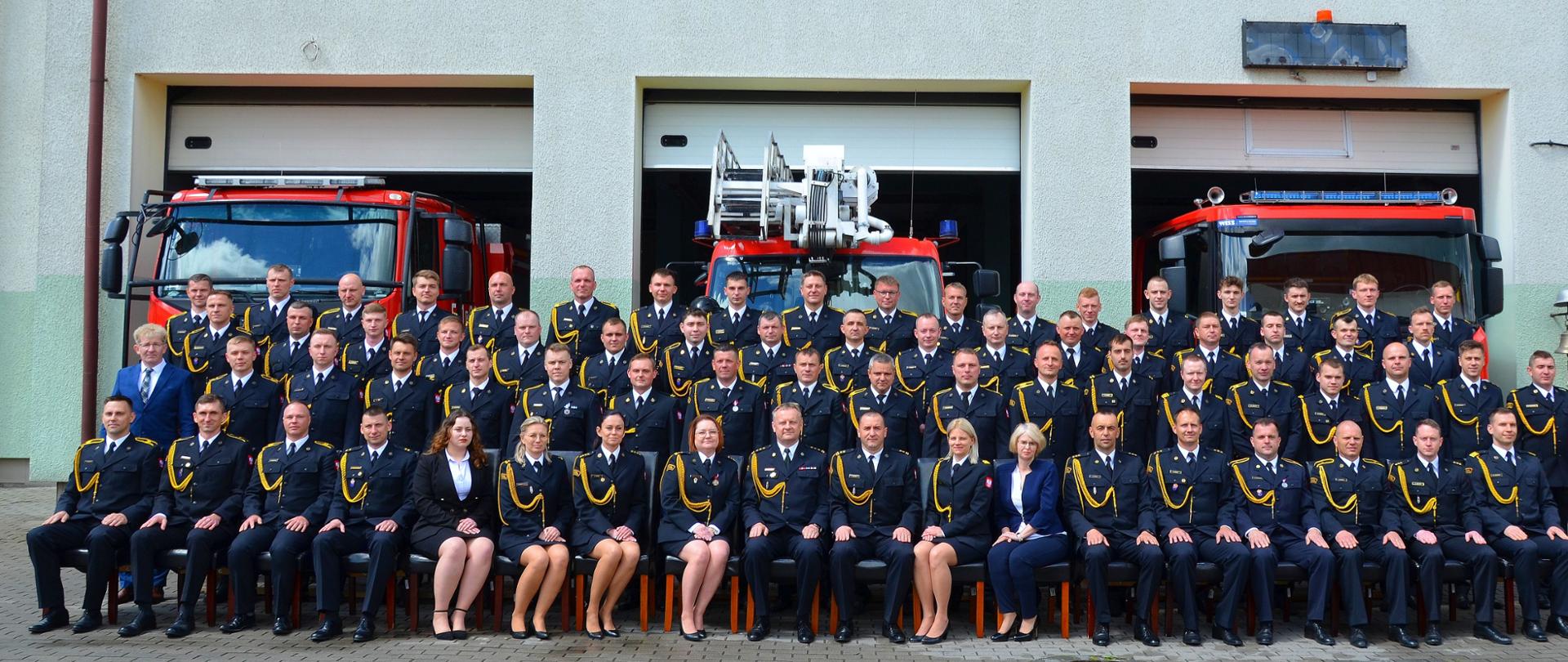 Na zdjęciu wszyscy pracownicy Komendy ustawieni w trzech szeregach na tle garaży i samochodów pożarniczych, odświętne mundury bez czapek, w tle trzy samochody pożarnicze