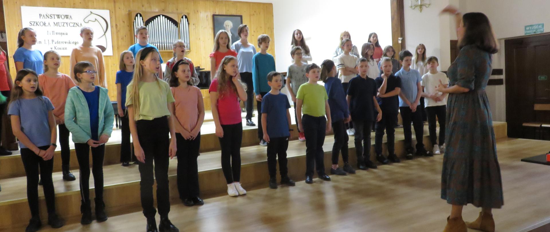 Chór stoi na scenie twarzą do publiczności, dyrygent przodem do nich