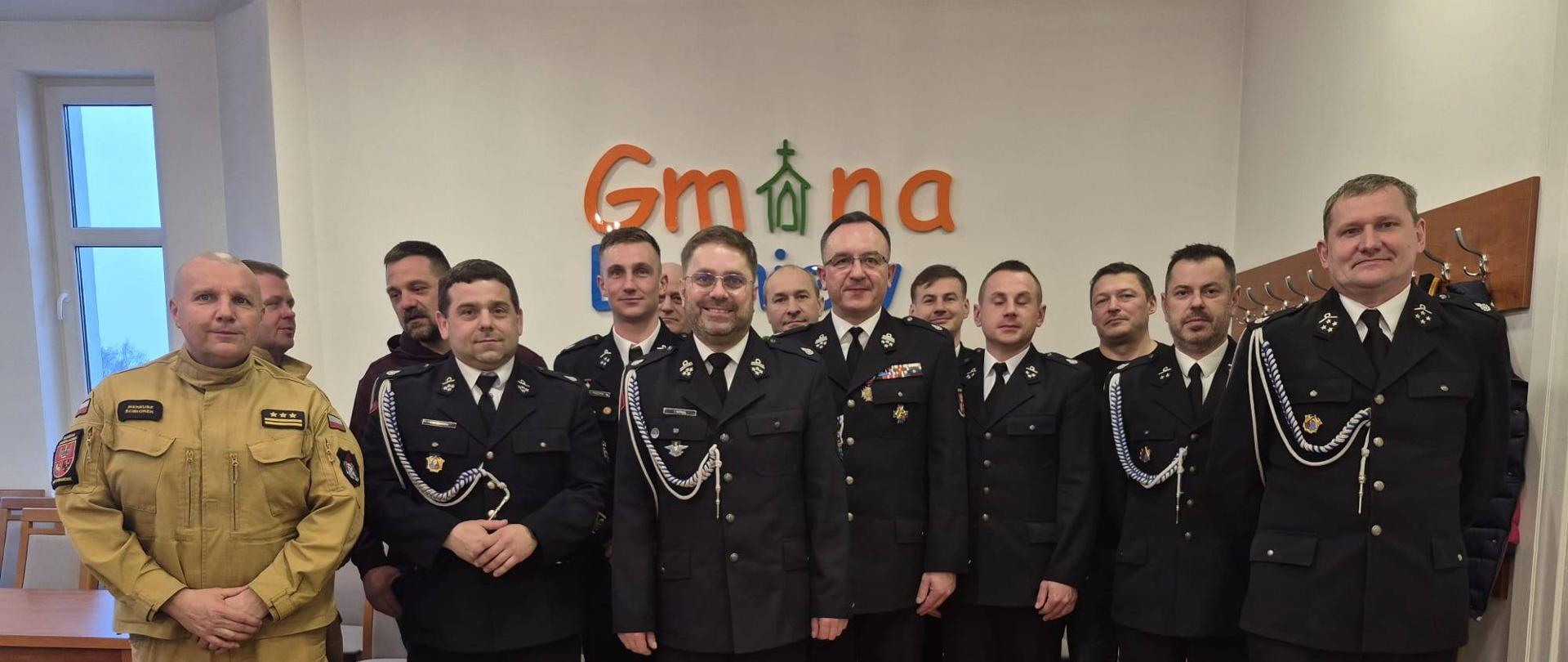 Do zdjęcia pozuje grupa strażaków ochotników w mundurach galowych. Z lewej strony strażak w jasnym mundurze.
