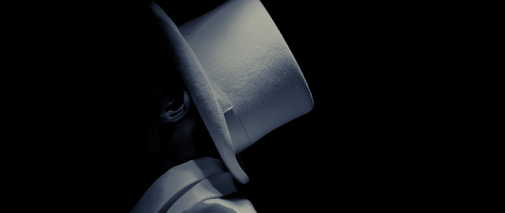 Mężczyzna w białym cylindrze i białym garniturze widziany z profilu na czarnym tle. Twarz lekko zarysowana, widoczne lewe ucho.