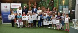 Na fotografii znajdują się organizatorzy i partnerzy konkursu wraz z grupą dzieci, które zajęły II miejsce w konkursie. Charakterystyczna jest „trawiasta” wyściółka, a w tle stoją wypchane zwierzęta z wystawy „Przyroda północno-wschodniej Polski” takie jak m.in. żubr i niedźwiedź. 