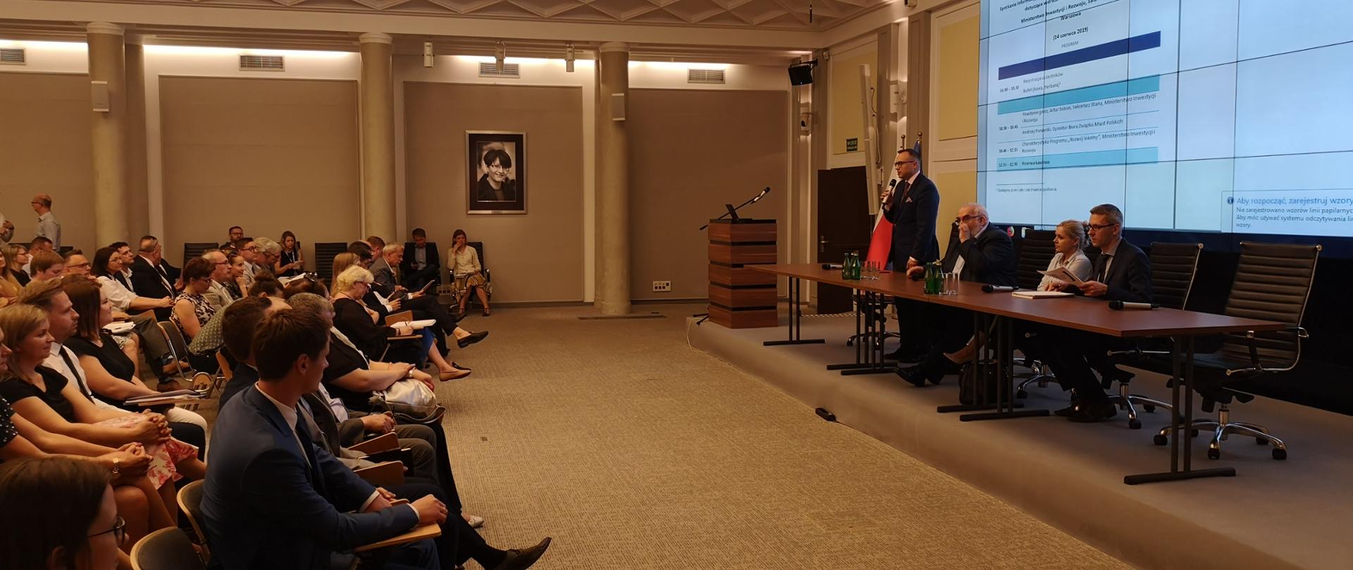 Po lewej stronie część osób na widowni, po prawej podest, na podeście długi stół, z tyłu ekran z agendą wydarzenia. Przy stole siedzą cztery osoby, pierwszy z lewej wiceminister Artur Soboń.