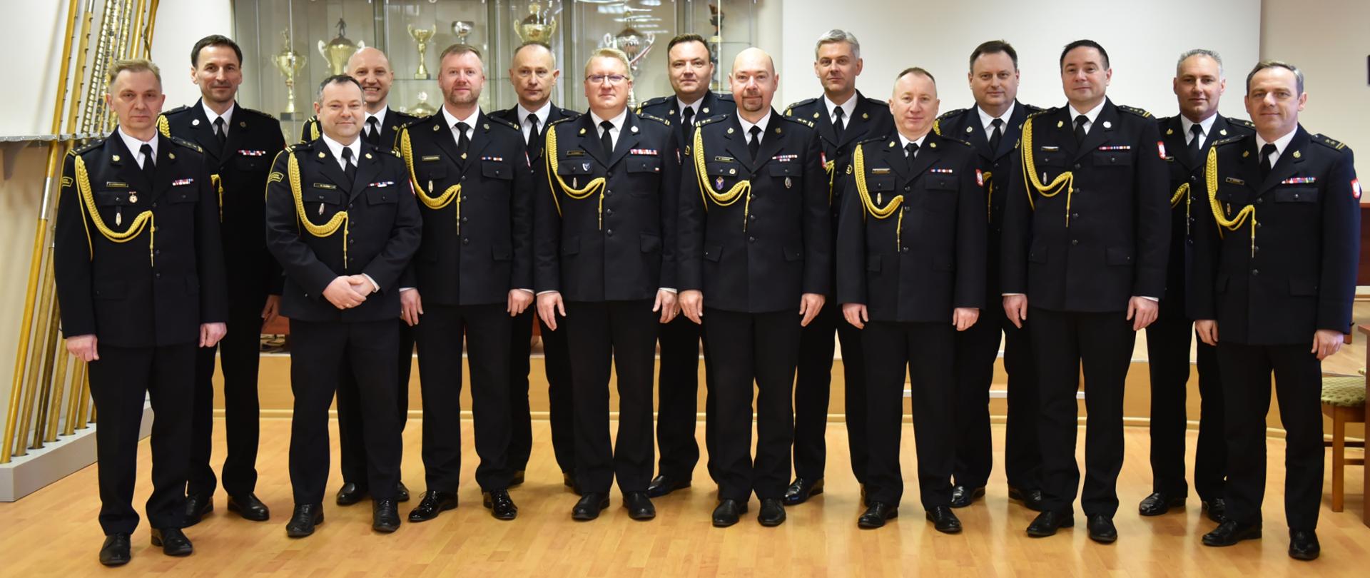 na zdjęciu piętnastu strażaków w mundurach wyjściowych stoją w dwóch rzędach tak aby twarze wszystkich były widoczne, zdjęcie zrobione w sali, w tle drzewce sztandarów przeszklony regał z pucharami i biały ekran do rzutnika