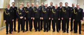 na zdjęciu piętnastu strażaków w mundurach wyjściowych stoją w dwóch rzędach tak aby twarze wszystkich były widoczne, zdjęcie zrobione w sali, w tle drzewce sztandarów przeszklony regał z pucharami i biały ekran do rzutnika