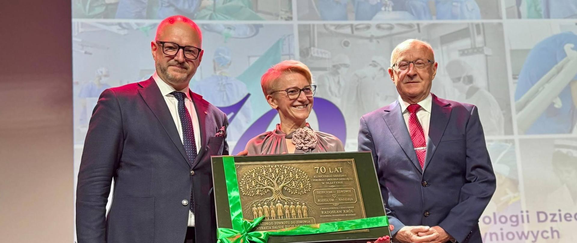Dwaj mężczyźni i kobieta stoją obok tablicy pamiątkowej, uśmiechając się do aparatu.
