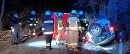 Na zdjęciu widać parawan przy którym stoją strażacy i policjanci a po prawej stronie przy drodze leży na dachu samochód terenowy. 