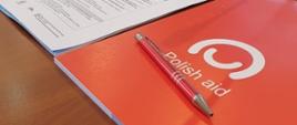Przestronna sala konferencyjna. Na drewnianym stole widoczne są mikrofony, słuchawki i materiały szkoleniowe - agenda, długopis i notes z logotypem Polskiej pomocy w języku angielskim.