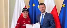 Minister Bortniczuk i żużlowiec Szymon Woźniak pozują do zdjęcia na tle flag Polski i Unii Europejskiej. Minister ubrany jest w granatowy garnitur w kratkę a żużlowiec w czerwoną bluzę reprezentacyjną z orzełkiem na piersi.