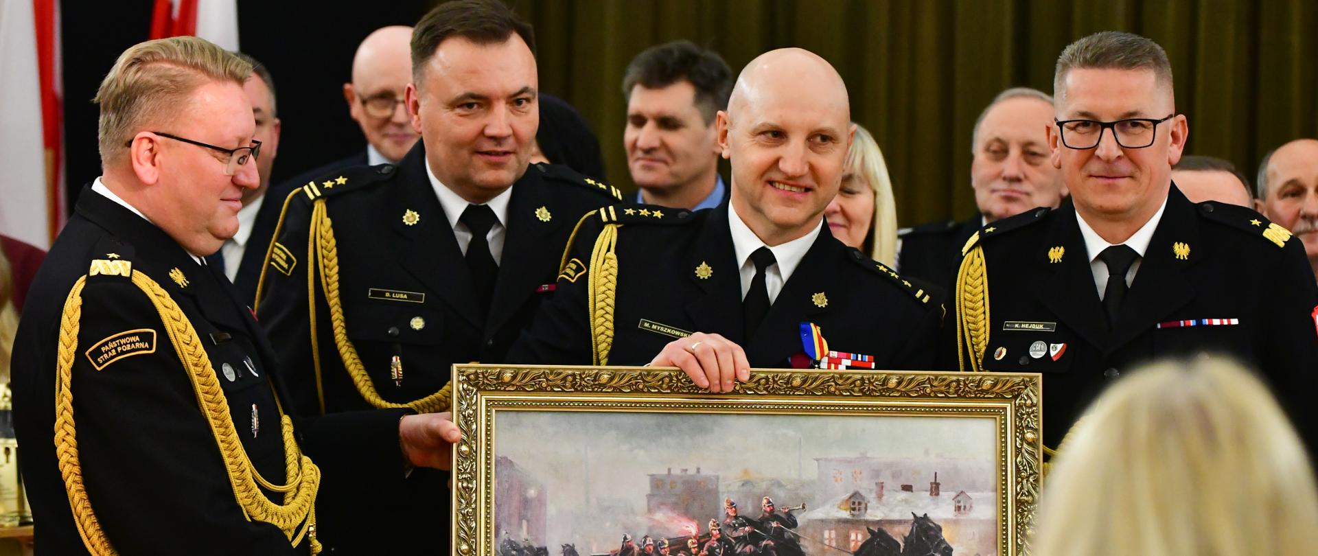 komendanci powiatowi PSP w kutnie, komendant wojewódzki PSP w łodzi oraz z-ca komendanta głównego PSP pozują do zdjecia trzymając obraz na którym strażacy wyjeżdżają do akcji wozem konnym