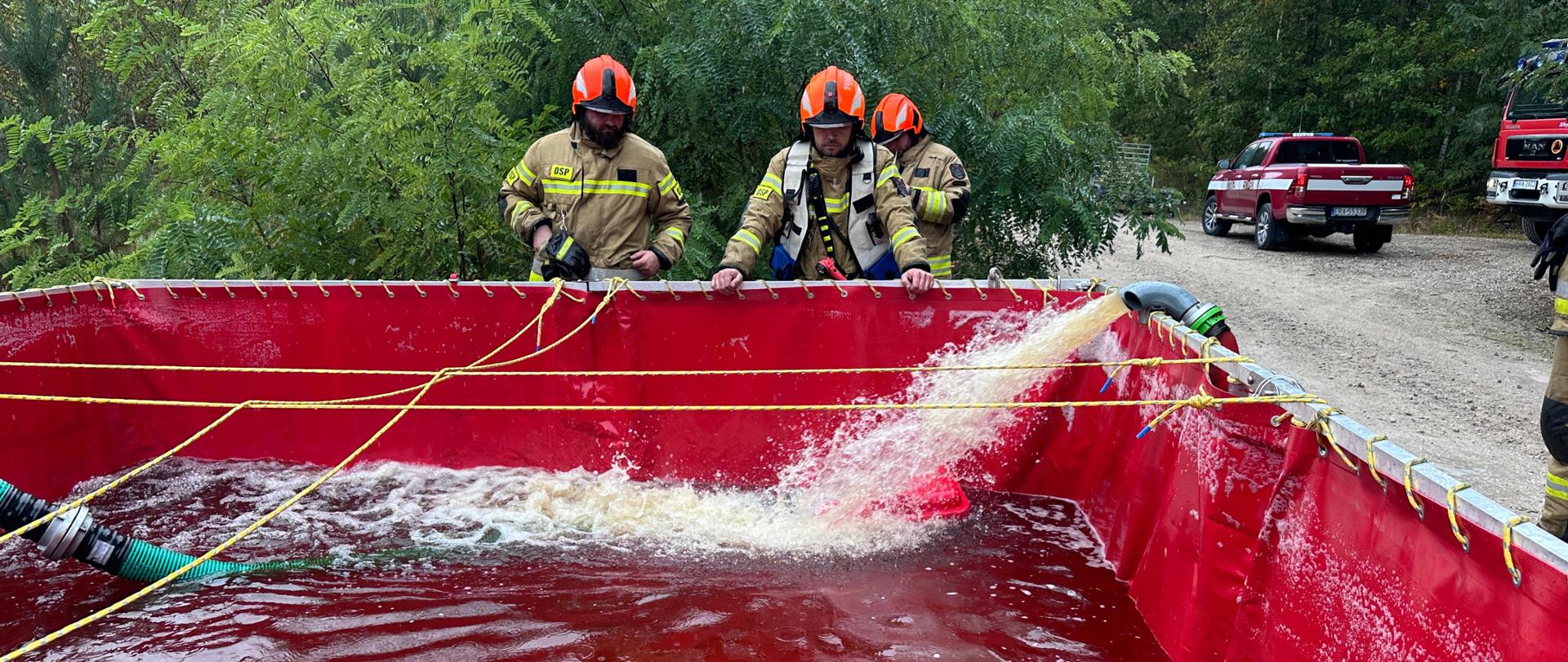 Ćwiczenia strażaków przy dużym czerwonym zbiorniku na wodę. Widać wypływającą wodę i kilku strażaków w kaskach pomarańczowych obserwujących napełnianie zbiornika. W tle leśna droga i samochód pożarniczy.