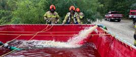 Ćwiczenia strażaków przy dużym czerwonym zbiorniku na wodę. Widać wypływającą wodę i kilku strażaków w kaskach pomarańczowych obserwujących napełnianie zbiornika. W tle leśna droga i samochód pożarniczy.