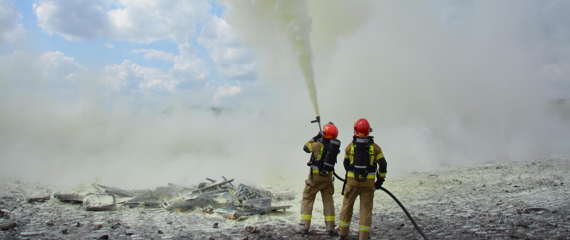 Dwóch strażaków ubranych w ubrania specjalne, hełmy oraz aparaty powietrzne stoi obok pogorzeliska. Trzymają odcinek z prądownicą, skierowaną ku górze z której wydobywa się proszek. W tle widoczne obłoki chmur.