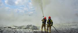 Dwóch strażaków ubranych w ubrania specjalne, hełmy oraz aparaty powietrzne stoi obok pogorzeliska. Trzymają odcinek z prądownicą, skierowaną ku górze z której wydobywa się proszek. W tle widoczne obłoki chmur.