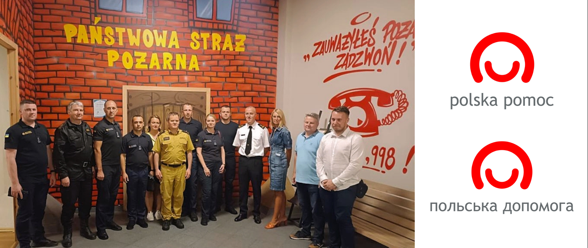 Polscy i ukraińscy strażacy pozują do grupowego zdjęcia w sali edukacyjnej, nad grupą na ścianie napis PAŃSTWOWA STRAŻ POŻARNA, po prawo na ścianie wizerunek telefonu i cyfry 998 oraz napis "Zauważyłeś pożar, zadzwoń!"