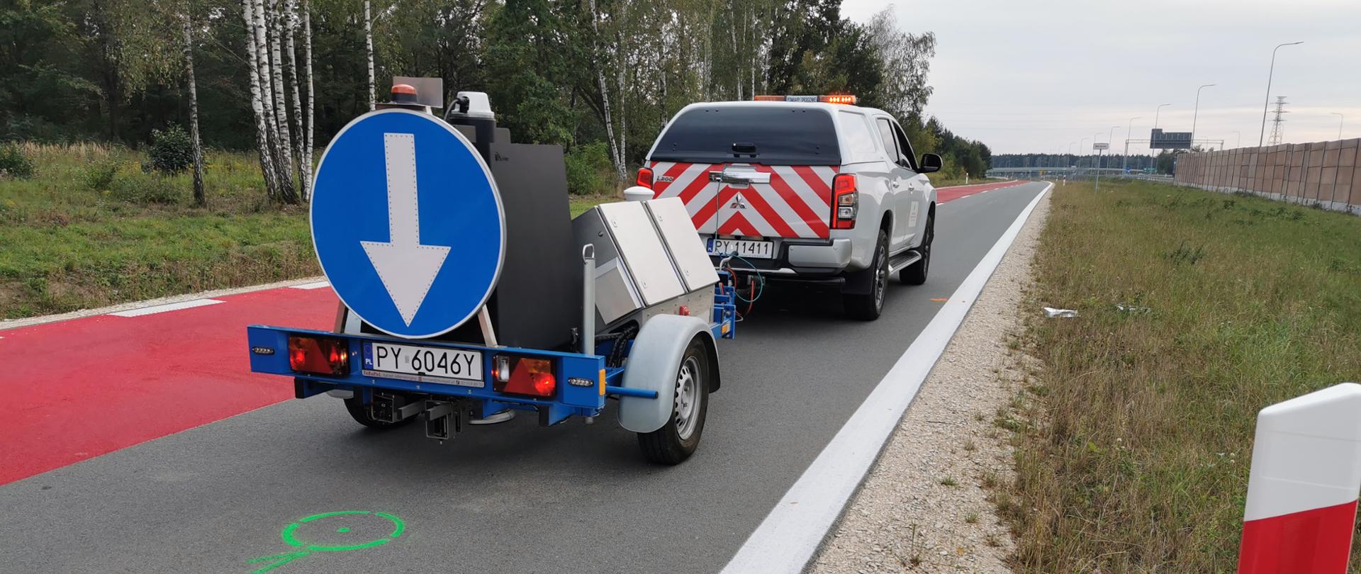 Samochód techniczny ciągnący przyczepę wyposażoną w aparaturę pomiarową wykonuje badania drogi. Na nawierzchni widoczne są kolorowe oznaczenia wykorzystywane w trakcie pomiarów i kontroli parametrów infrastruktury.