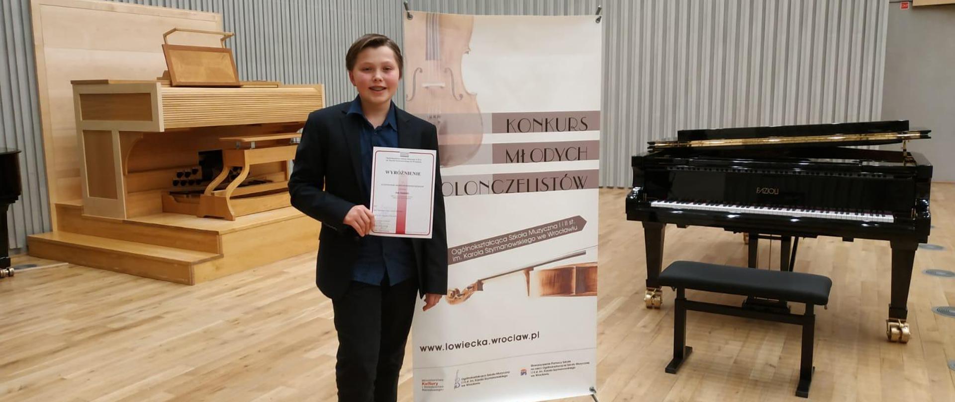 Jan Jasiński trzymający dyplom wyróżnienia na tle baneru konkursowego młodych wiolonczelistów