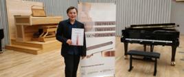 Jan Jasiński trzymający dyplom wyróżnienia na tle baneru konkursowego młodych wiolonczelistów