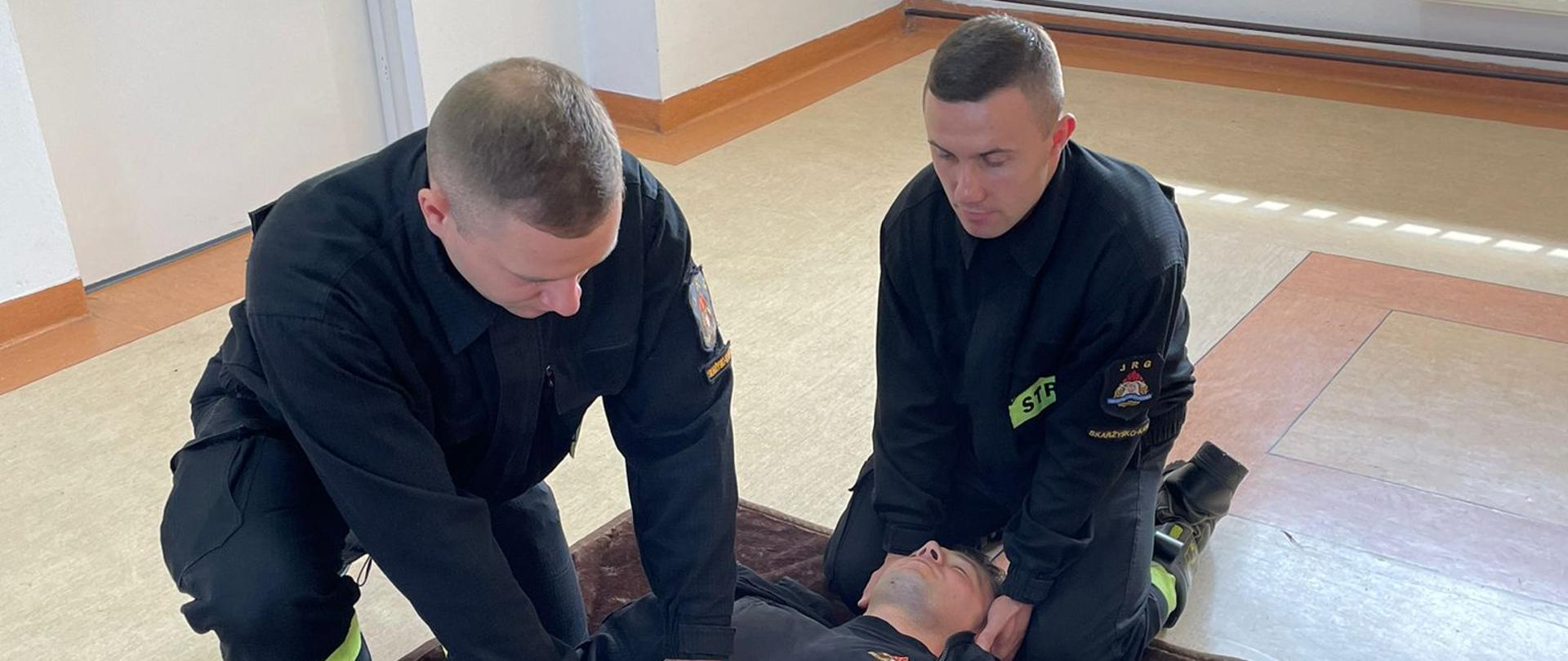 Na zdjęciu trzecim strażak-ratownik wykonuje symulację badania urazowego osoby poszkodowanej. Badanie rozpoczyna od głowy, następnie bada szyję, obojczyki, klatkę piersiową, brzuch, miednicę, kończyny dolne, kończyny górne oraz plecy i pośladki. Badanie urazowe polega na szybkim zidentyfikowaniu krwawień oraz innych nieprawidłowości poszczególnych części ciała.