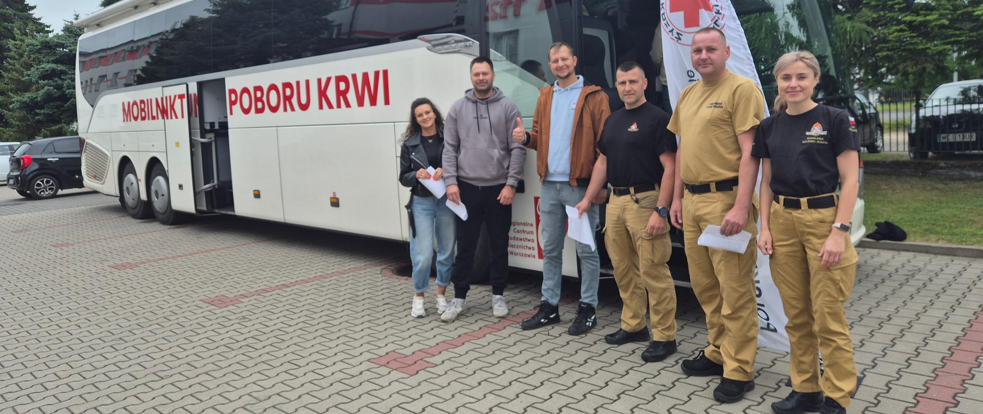 Grupa osób czekająca przed autobusem krwiodawstwa. Troje ubranych w umundurowanie służbowe oraz troje w ubrania prywatne. Autobus stoi przed budynkiem komendy PSP na parkingu 
