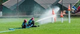 Dwóch strażaków OSP w czarnych ubraniach koszarowych bierze udział w zawodach sportowo-pożarniczych. Na murawie stadionu sportowego trzymają odcinek węża i strumieniem wody przewracają ustawione na podstawach stożki ostrzegawcze