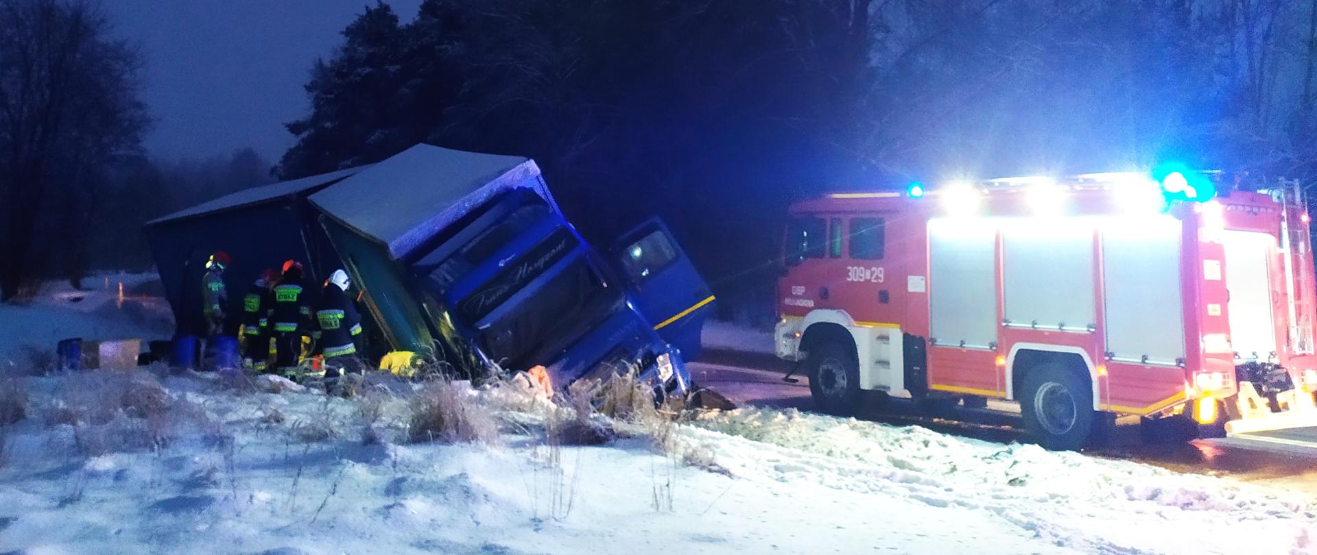 Zdjęcie przedstawia samochód ciężarowy w rowie z uszkodzoną kabiną. Wokół ciężarówki stoją strażacy. Przed samochodem ciężarowym stoi samochód strażacki oświetlający teren akcji. Na zewnątrz panuje półmrok.