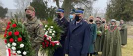 Dwie osoby delegacji PSP w płaszczach granatowych umundurowania wyjściowego. Strażak z lewej strony trzyma wiązankę kwiatów biało-czerwonych. Przed nimi stoi Żołnierz Wojska Polskiego również trzymający wiązankę biało-czerwonych kwiatów. W tle kilku harcerzy w płaszczach koloru zielonego. Wszystkie osoby na zdjęciu mają maseczki na twarzy, 