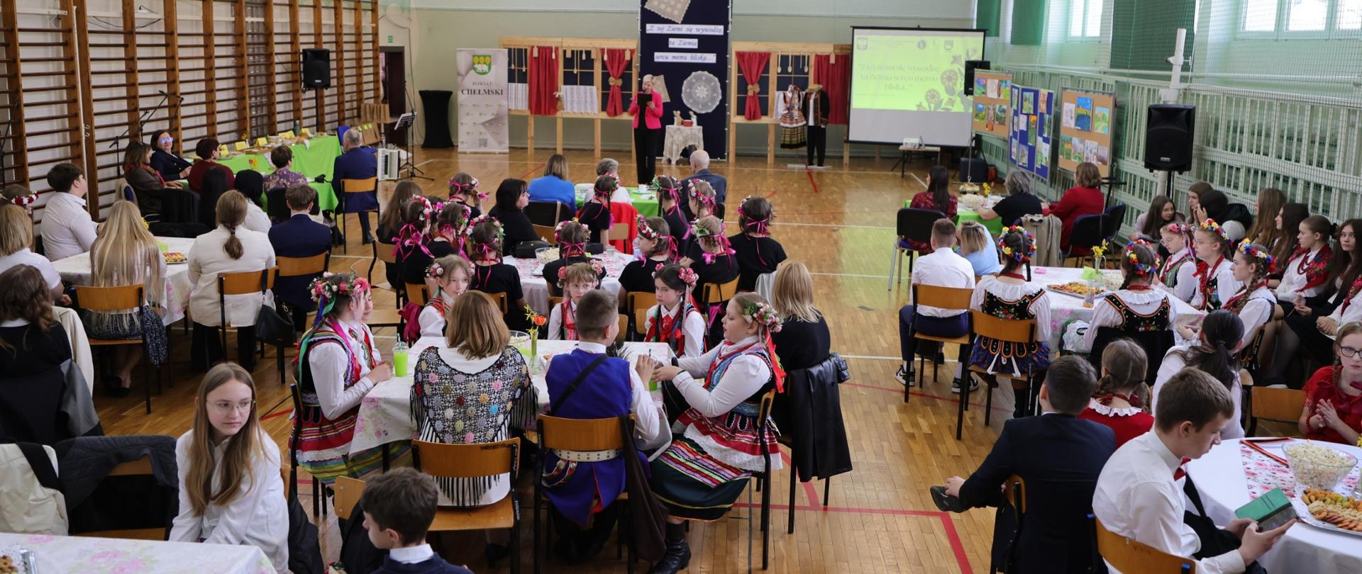 Uczestnicy wydarzenia siedzą przy stołach w sali gimnastycznej podczas przeglądu artystycznego.