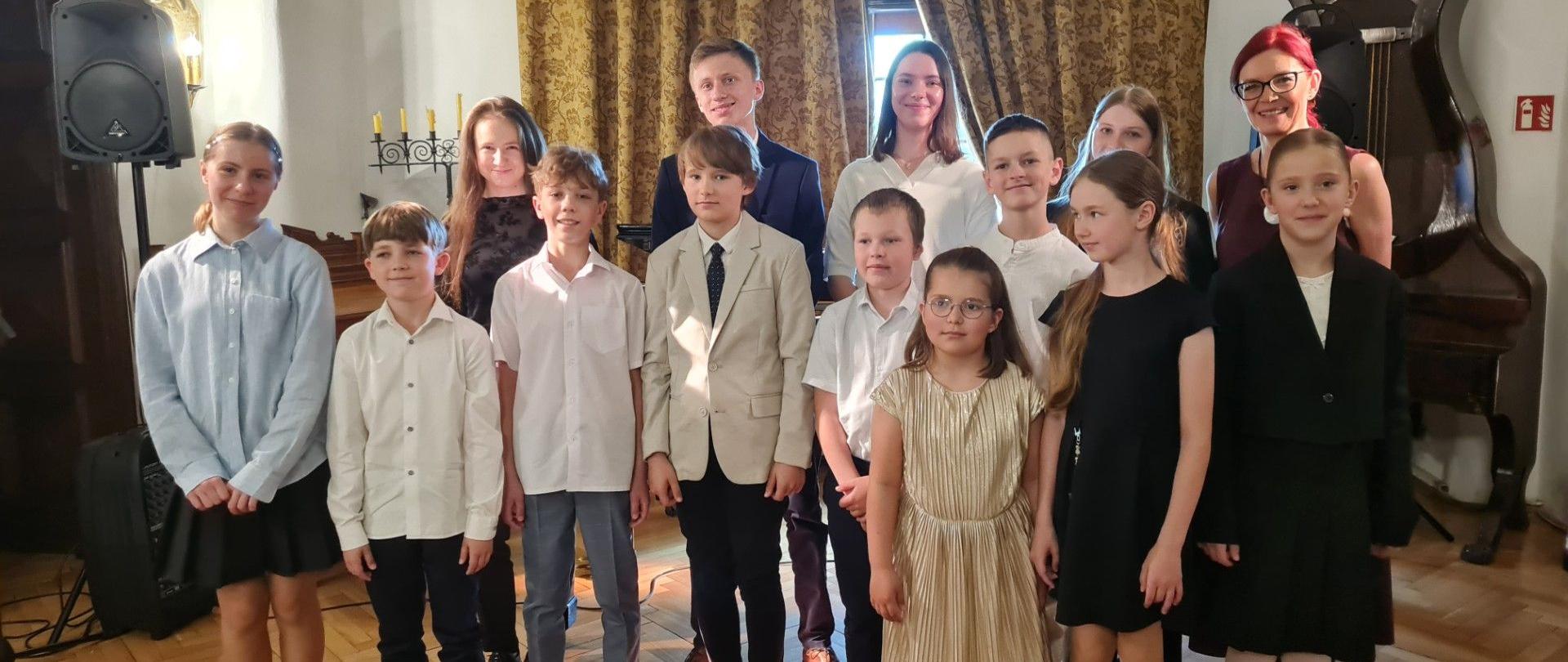Grupa dzieci i młodzieży pozuje do wspólnego zdjęcia w formalnych strojach. W przednim rzędzie stoją dzieci ubrane w białe koszule, a jeden chłopiec ma na sobie beżowy garnitur i krawat. Po ich prawej stronie stoi dziewczynka w błyszczącej złotej sukience. W tylnym rzędzie widoczni są starsi uczniowie oraz dwie dorosłe kobiety – jedna w białej bluzce, a druga z rudymi włosami, w ciemnym topie i okularach. W tle widać bogato zdobione zasłony, kandelabr oraz fragment instrumentu muzycznego.
