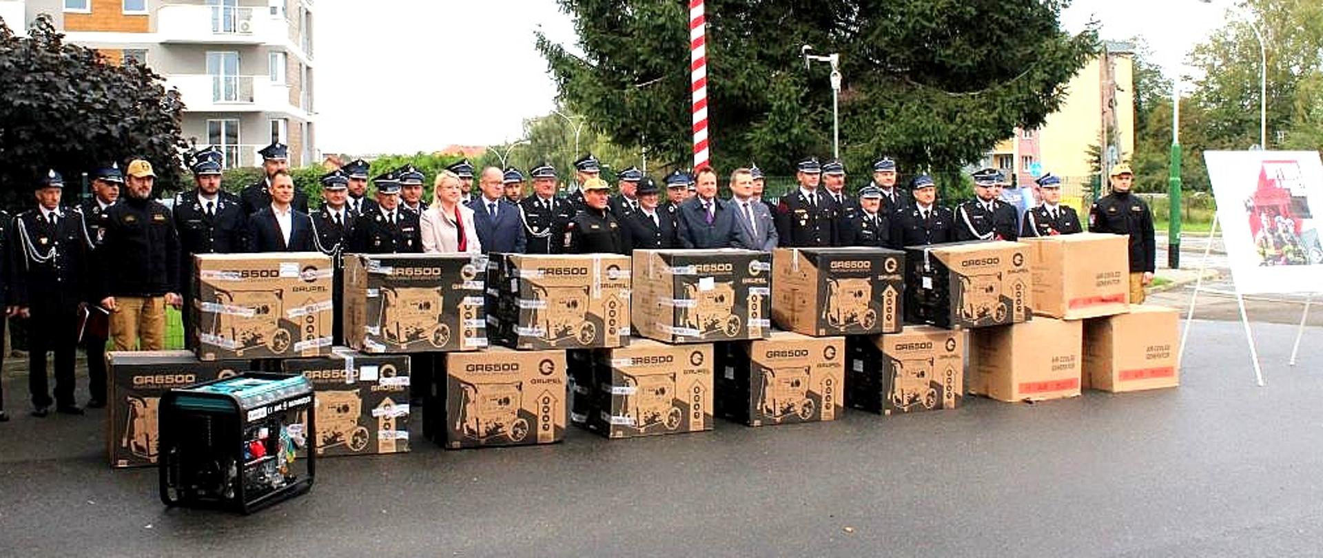 Zdjęcie zrobione na zewnątrz, na placu przed komendą straży pożarnej. Zaproszeni goście stoją za ustawionymi agregatami prądotwórczymi