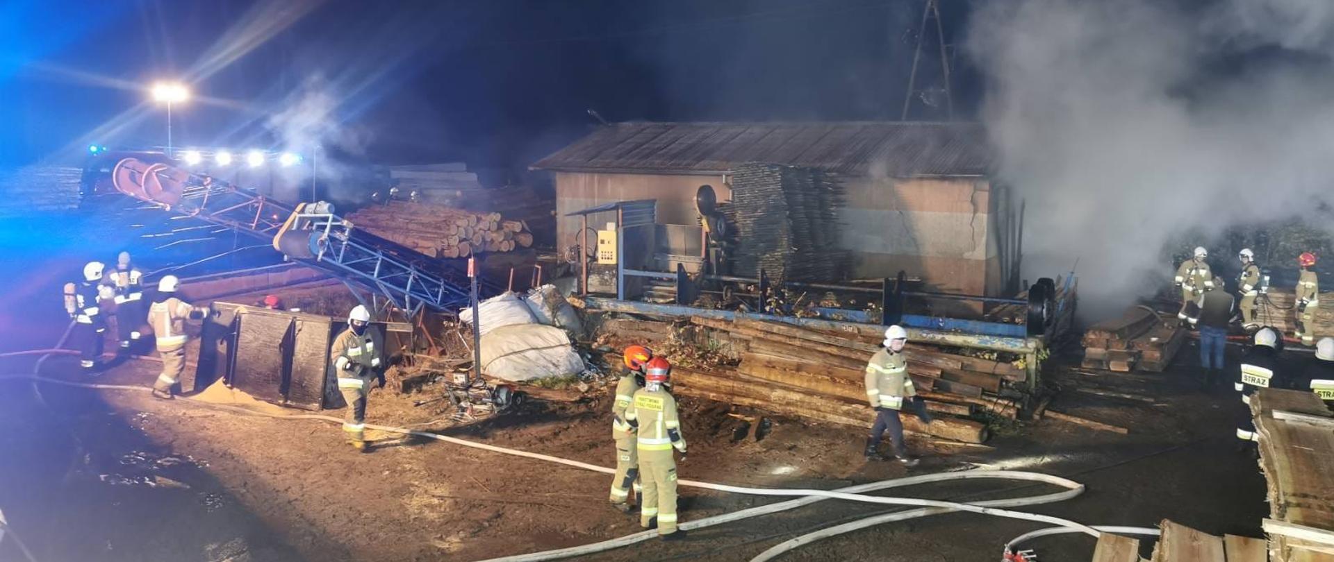 Objęty pożarem budynek, z którego unoszą się kłęby dymu. Przed budynkiem znajdują się strażacy prowadzący działania gaśnicze.