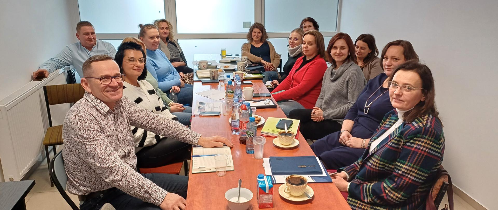 Na zdjęciu widać grupę osób siedzących przy stole w jasnym biurowym pomieszczeniu. Na stole znajdują się dokumenty i teczki. 