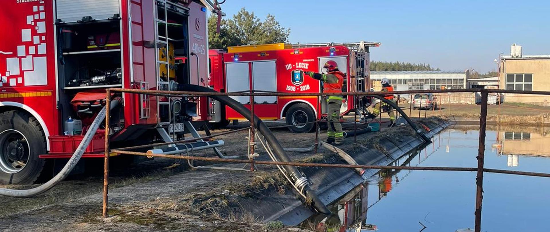 Zdjęcie przedstawia strażaków uczestniczących w ćwiczeniach na terenie zakładu meblarskiego.
W tle samochody strażackie.
