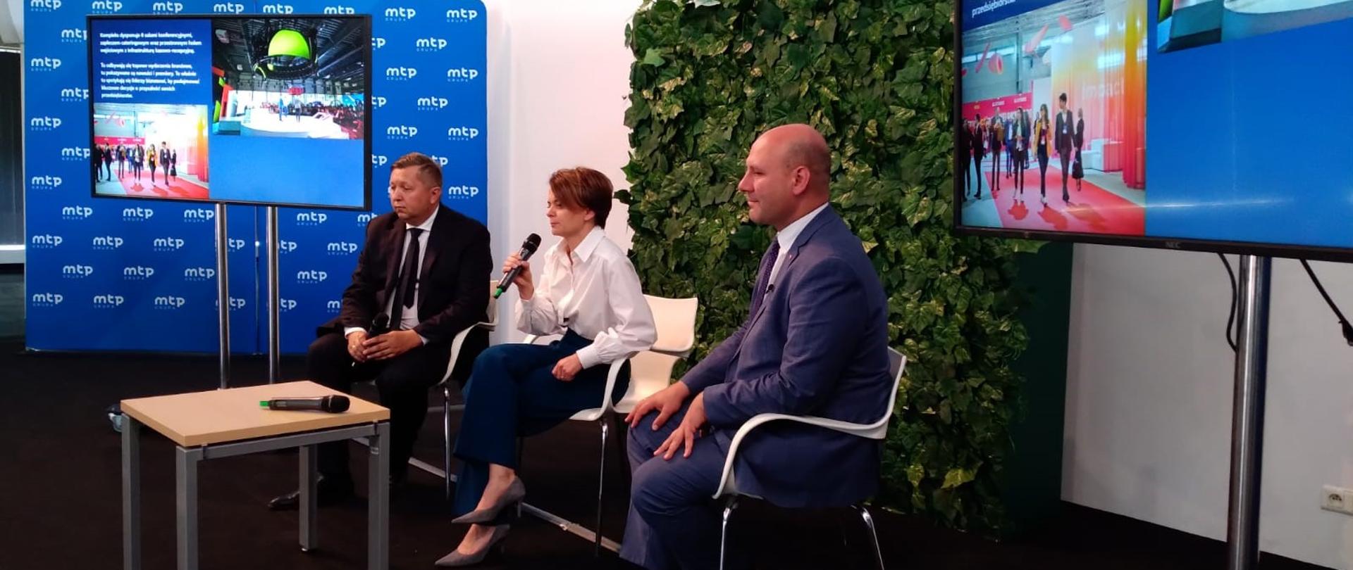 Trzy osoby siedzą obok siebie na scenie. W środku z mikrofonem w prawej dłoni siedzi wiceminister Jadwiga Emilewicz. 