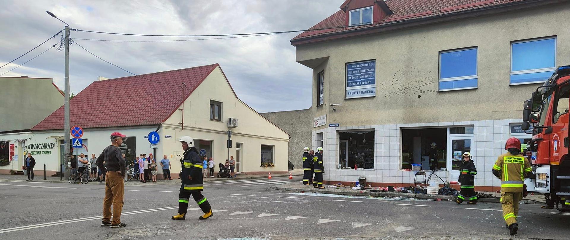 Zdjęcie przedstawia budynek narożny, w którym doszło do wybuchu. W budynku wypadły szyby w oknach. Na zewnątrz strażacy wykonujący zadania ratownicze oraz będący świadkami zdarzenia.