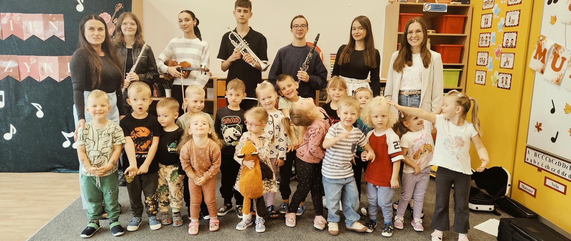 Grupa przedszkolaków oraz starszych uczniów z instrumentami muzycznymi pozuje wspólnie do zdjęcia w sali przedszkola podczas audycji muzycznej z okazji Międzynarodowego Dnia Muzyki.
