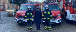 Na zdjęciu Komendant Powiatowy PSP w Myśliborzu ubrany w granatowy płaszcz służbowy i rogatywkę. Obok stoją dwaj strażacy - ochotnicy ubrani w umundurowanie bojowe z hełmem. Panowie stoją na tle pojazdów pożarniczych pożarniczych.
