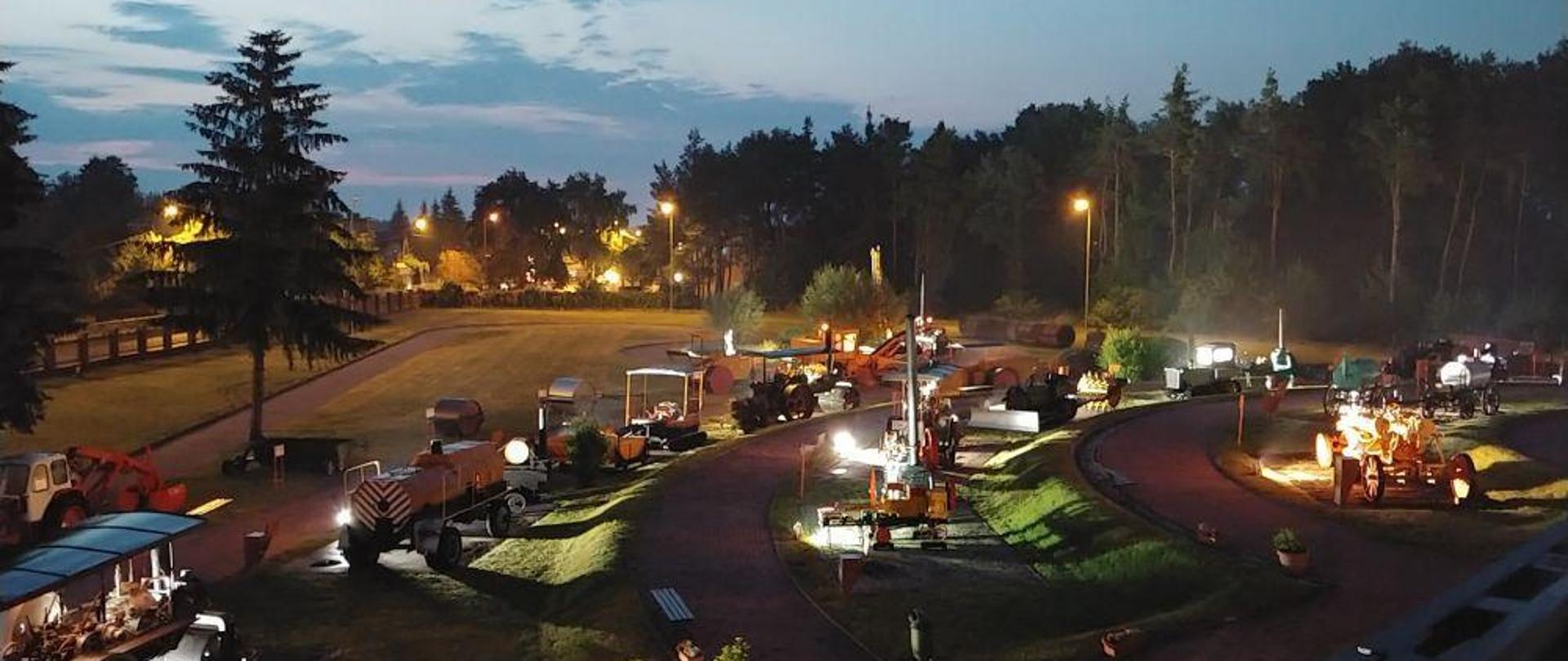 Nocne zdjęcie terenu wokół budynku muzeum. Widoczne eksponaty muzealne, drzewa i oświetlenie z lamp