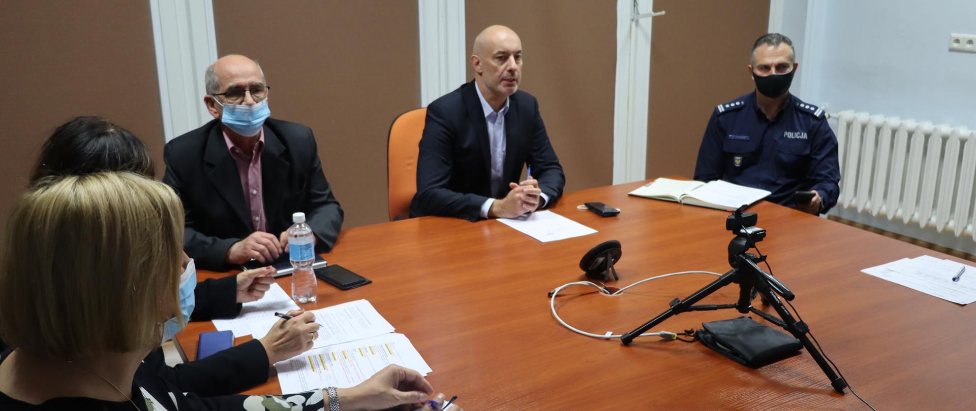 Na środku kadru wojewoda opolski przemawiający do zgromadzonych na widekonferencji. Obok wojewody, za stołem, zasiada dyrektor wydziału bezpieczeństwa oraz - po prawej stronie od wojewody, zastępca opolskiego komendanta wojewódzkiej policji. Na stole umieszczona kamera oraz dokumenty. W tle rolety w oknach. 
