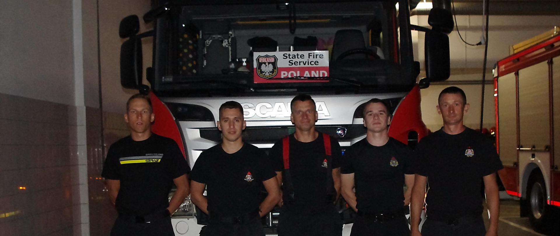 Pięciu strażaków ubranych w mundury koszarowe stoi przed przedem ciężakiego samochodu gaśniczego, za którego szybą jest widoczna tabliczka z napisem state fire service Poland