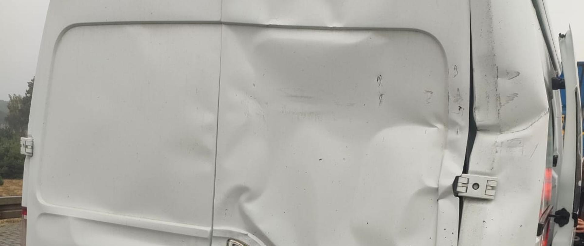 Tył białego vana marki Renault Master z widocznymi uszkodzeniami po kolizji. Tylne drzwi są mocno wgniecione i zdeformowane, szczególnie po lewej stronie. Prawa tylna lampa jest przekrzywiona, choć działa. Zderzak tylny jest uszkodzony i zarysowany, szczególnie po prawej stronie. Drzwi są nieszczelne i źle się domykają, co sugeruje uszkodzenie zawiasów lub ramy pojazdu. Zdjęcie wykonane na mokrej nawierzchni drogi przy pochmurnej pogodzie.