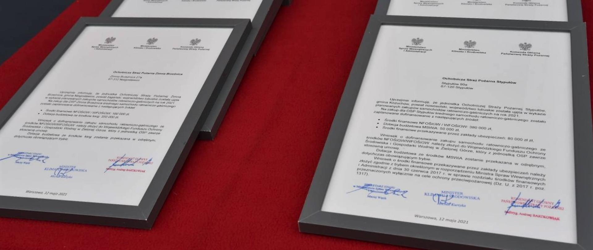 Promesy oprawione w stalowoszare ramki leżą na stole przykrytym czerwonym płótnem 