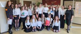 Na zdjęciu grupa uczniów. Wszyscy ubrani na galowo (czarne spodnie lub spódnice i białe bluzki). Koło uczniów po prawej stronie zdjęcia kobieta- dyrygent w czarnej bluzce i spódnicy, a po prawej kobieta- akompaniator w fioletowej sukience i czarnej marynarce. Tło obrazka w odcieniach beżu. 