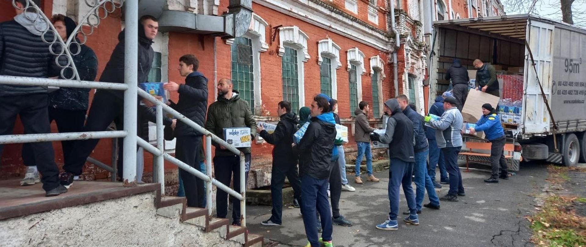 Przed budynkiem grupa mężczyzn rozpakowuje samochód z pomocą humanitarną, przekazując sobie paczki z rąk do rąk