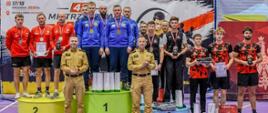 Uroczysta dekoracja najlepszych drużyn podczas 42. Mistrzostw Polski Strażaków w Tenisie Stołowym. Na najwyższym, zielonym stopniu podium stoi pięciu strażaków reprezentujących województwo podkarpackie, ubranych w niebieskie stroje sportowe, ze złotymi medalami na szyjach. Po obu stronach znajdują się drużyny, które zajęły drugie i trzecie miejsce. Przed podium stoją dwaj przedstawiciele Państwowej Straży Pożarnej w umundurowaniu służbowym. W tle widoczny jest baner mistrzostw.