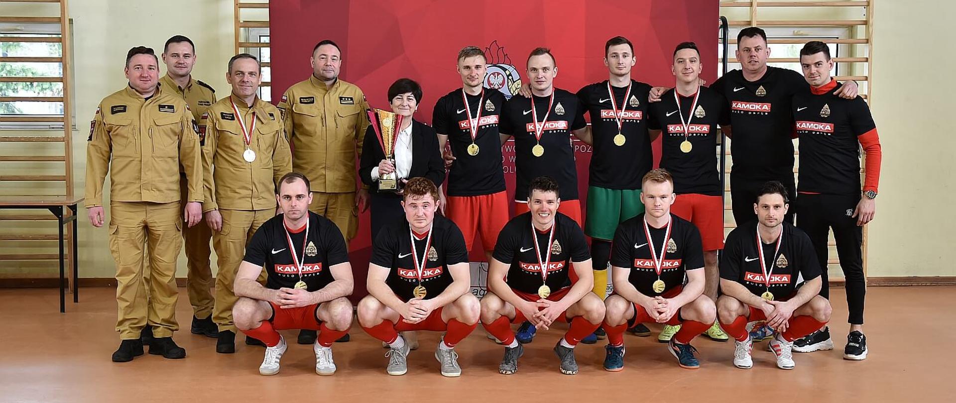 drużyna strażaków z Koluszek oraz komendanci pozują do zdjęcia na tle banneru